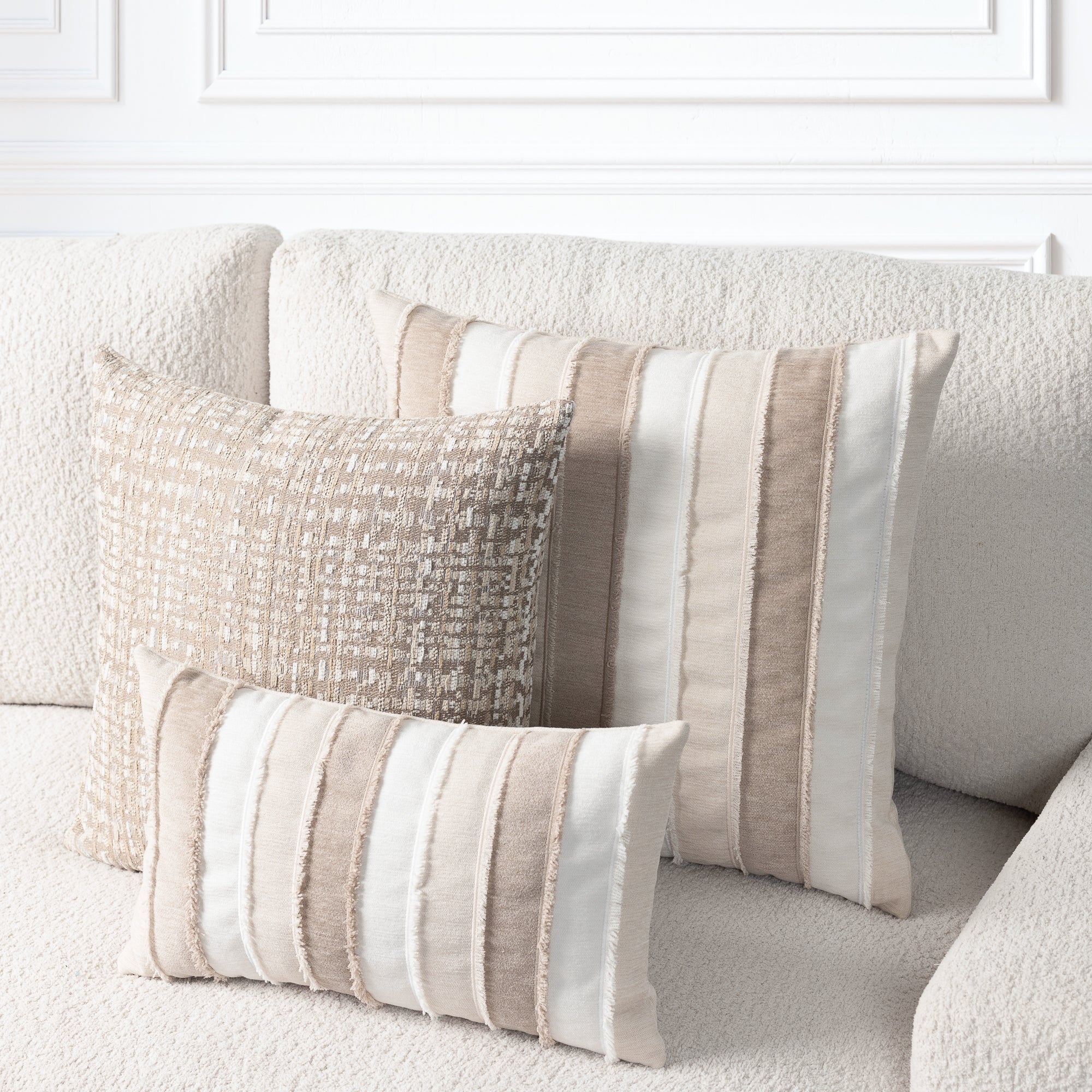 Three decorative throw pillows in neutral beige, cream, and taupe tones arranged on a textured cream sofa against a white paneled wall.