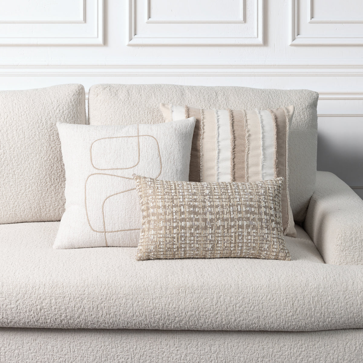 Three decorative throw pillows in neutral beige and cream tones arranged on a textured boucle sofa against a white paneled wall.