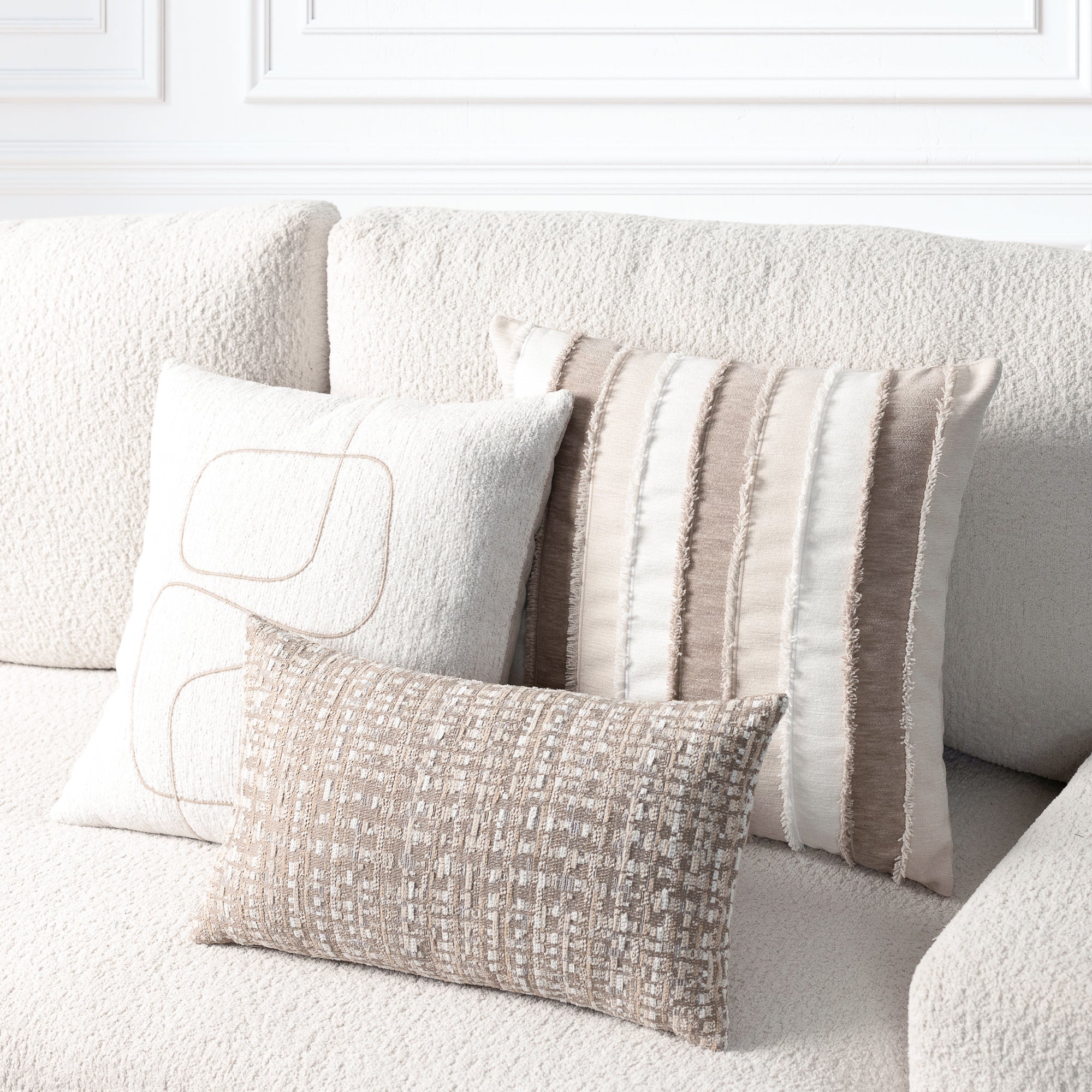 Three decorative throw pillows in neutral beige, cream, and taupe tones arranged on a textured cream sofa. One pillow features abstract stitched lines, another has vertical textured stripes, and the front pillow is woven with a basketweave pattern.