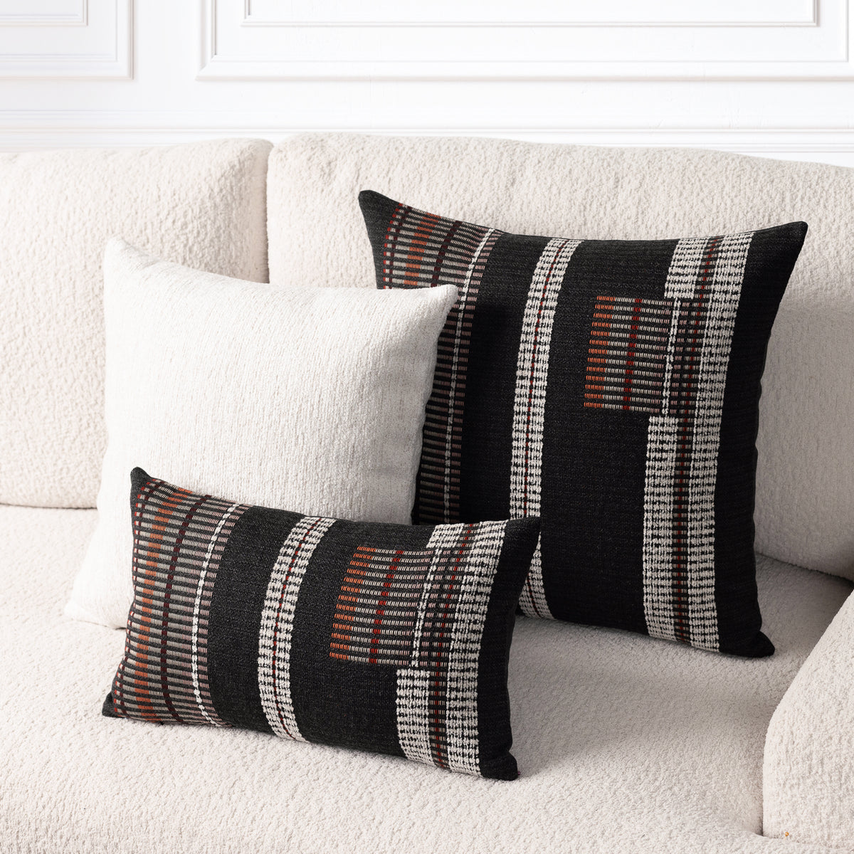 Two decorative throw pillows on a textured cream boucle sofa. One tall square pillow and one matching horizontal lumbar pillow, both featuring a black base with vertical white textured stripes and rust-orange geometric accents.